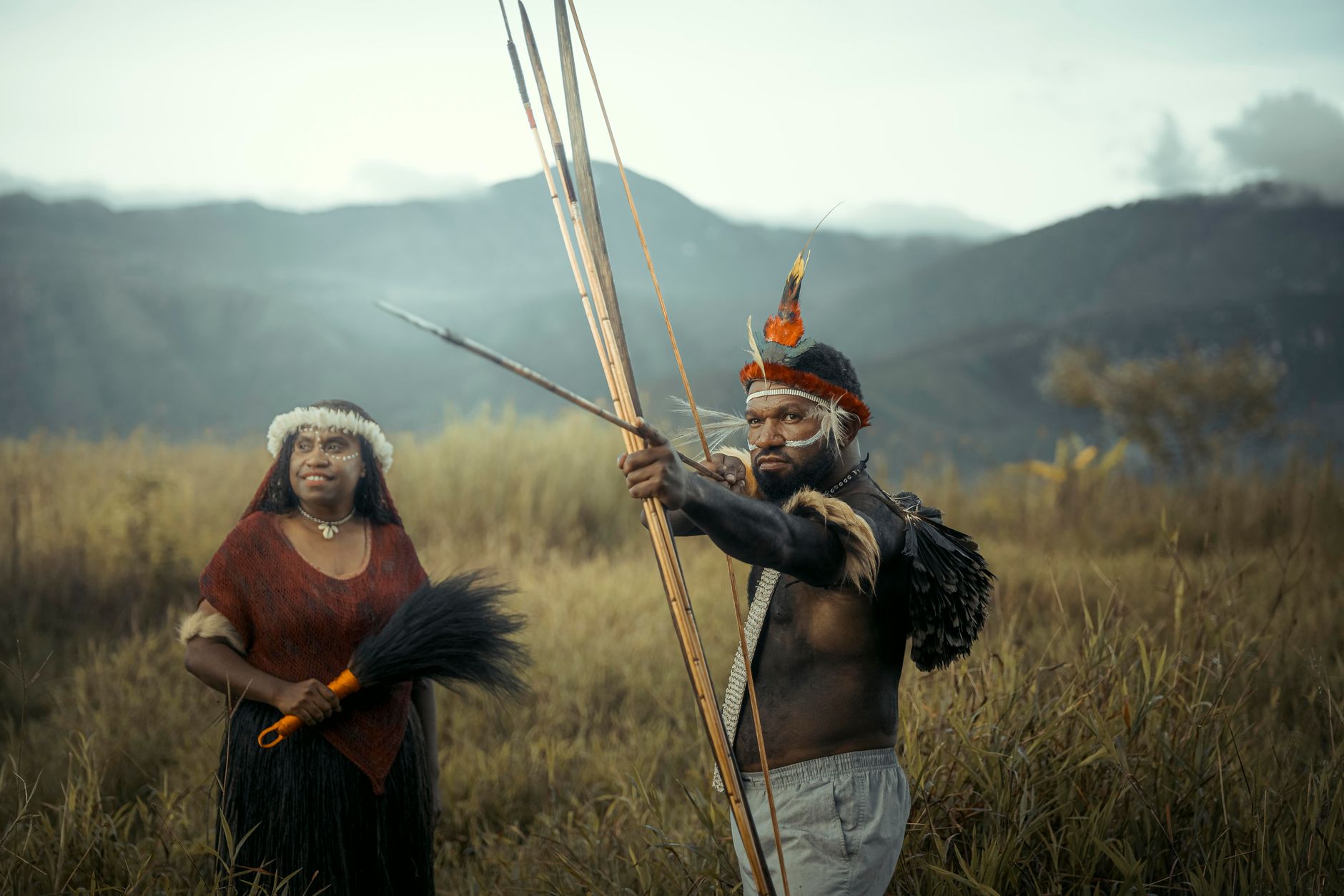 Portrait of indigenous people in traditional attire with bows and arrows set against a mountainous landscape.