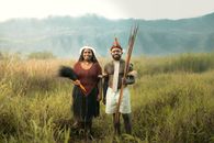 Couple in traditional dress stands in lush grassland with mountainous background.