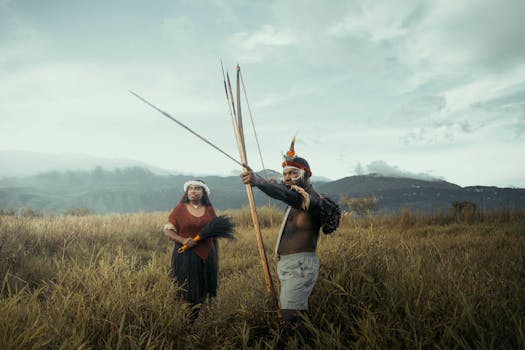 Indigenous duo holding traditional spears in a vast, green landscape with mountains.