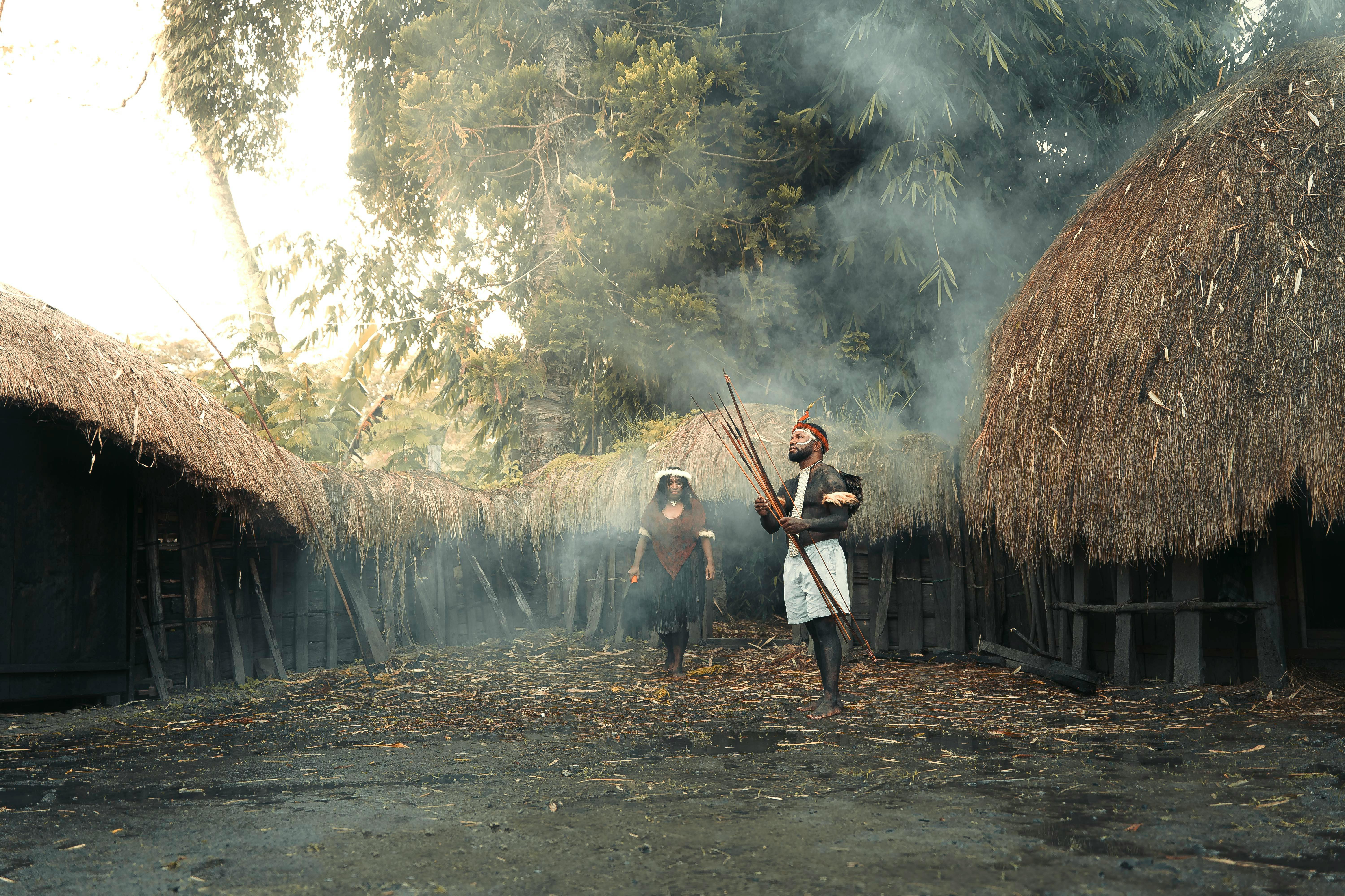 Explore a traditional indigenous village with tribal people in ceremonial attire amidst thatched huts and smoke.