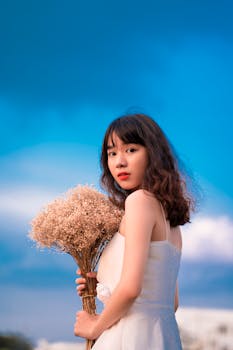一位年轻女子身着白色连衣裙，在蓝天的映衬下手持一束鲜花
