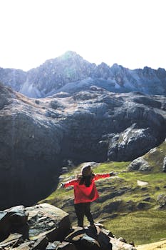 A woman in red stands on rocks, arms open to the majestic Huaral mountains under a bright sky.