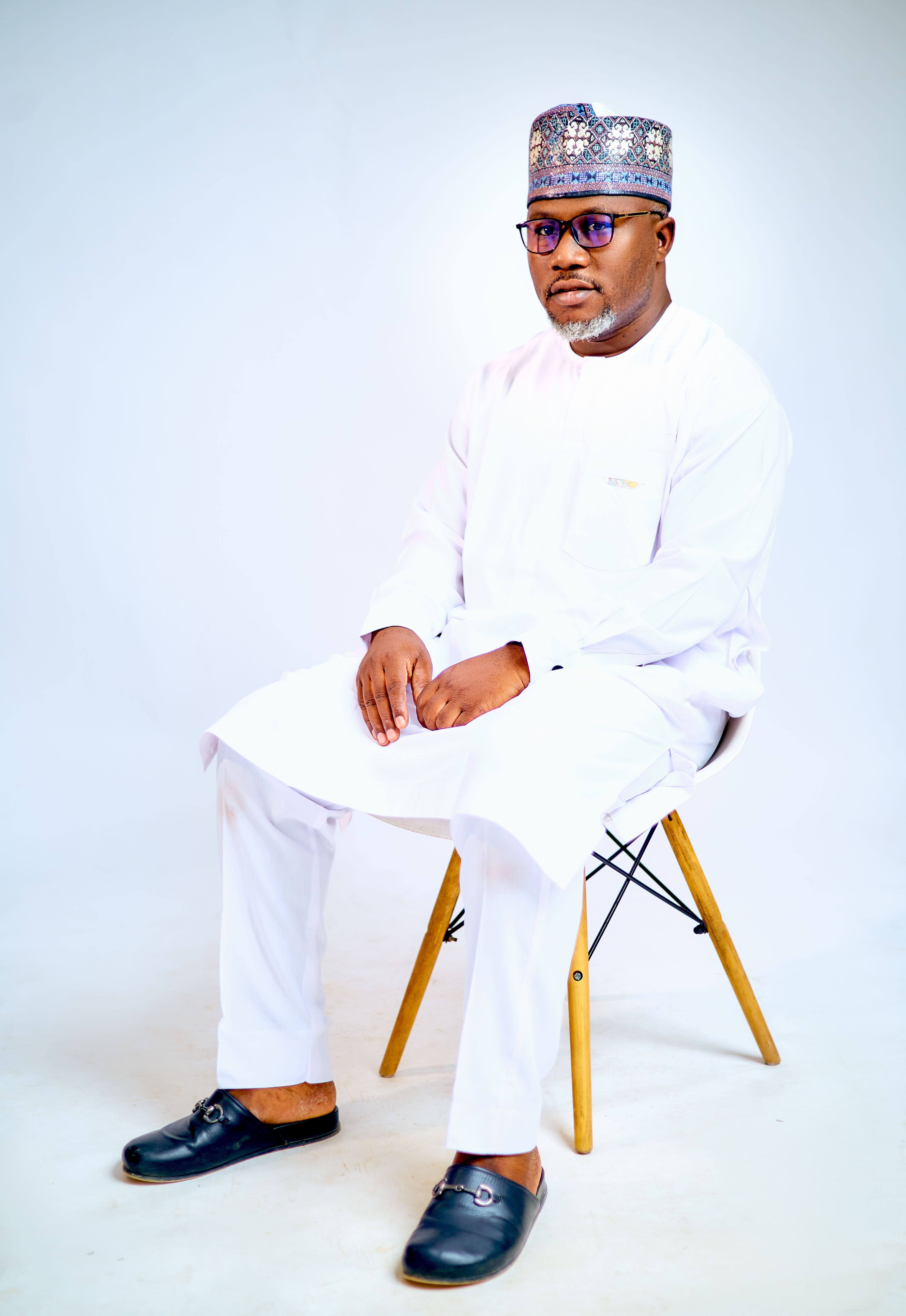 A man in traditional Nigerian clothing seated on a chair against a white backdrop.