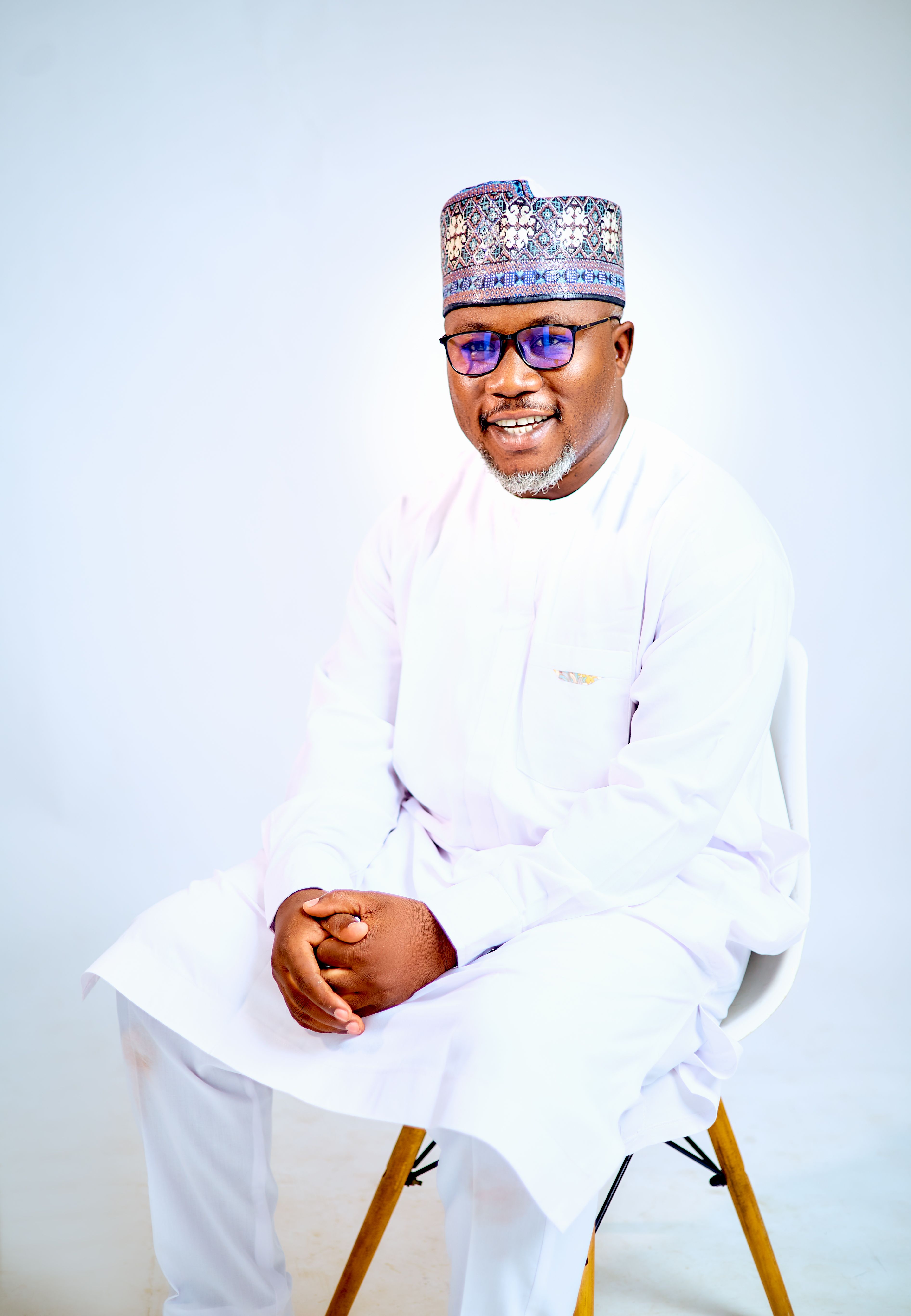 Portrait of a man in traditional white clothing and hat, sitting and smiling.