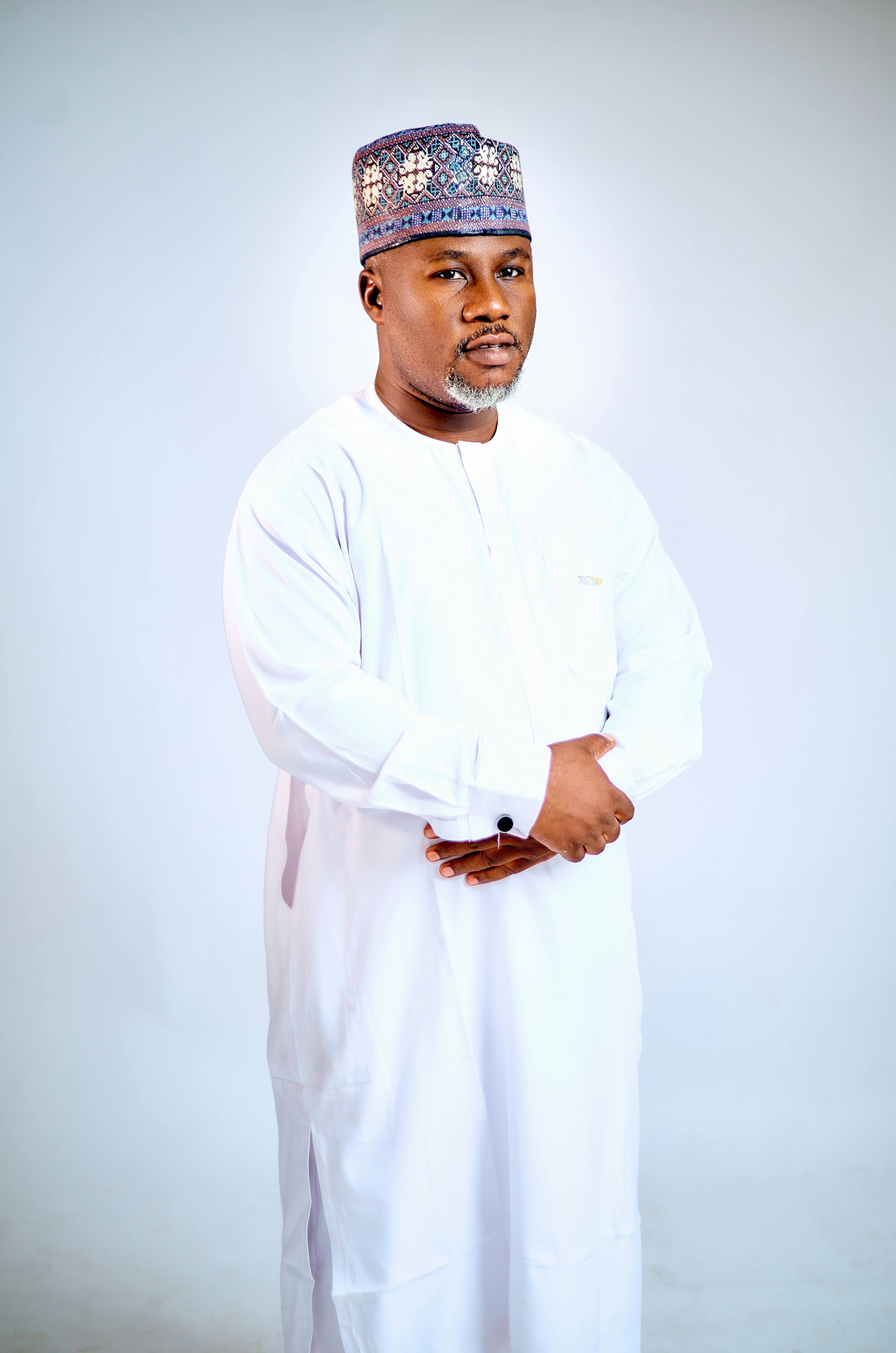 African man in white traditional attire with a patterned cap, standing confidently.