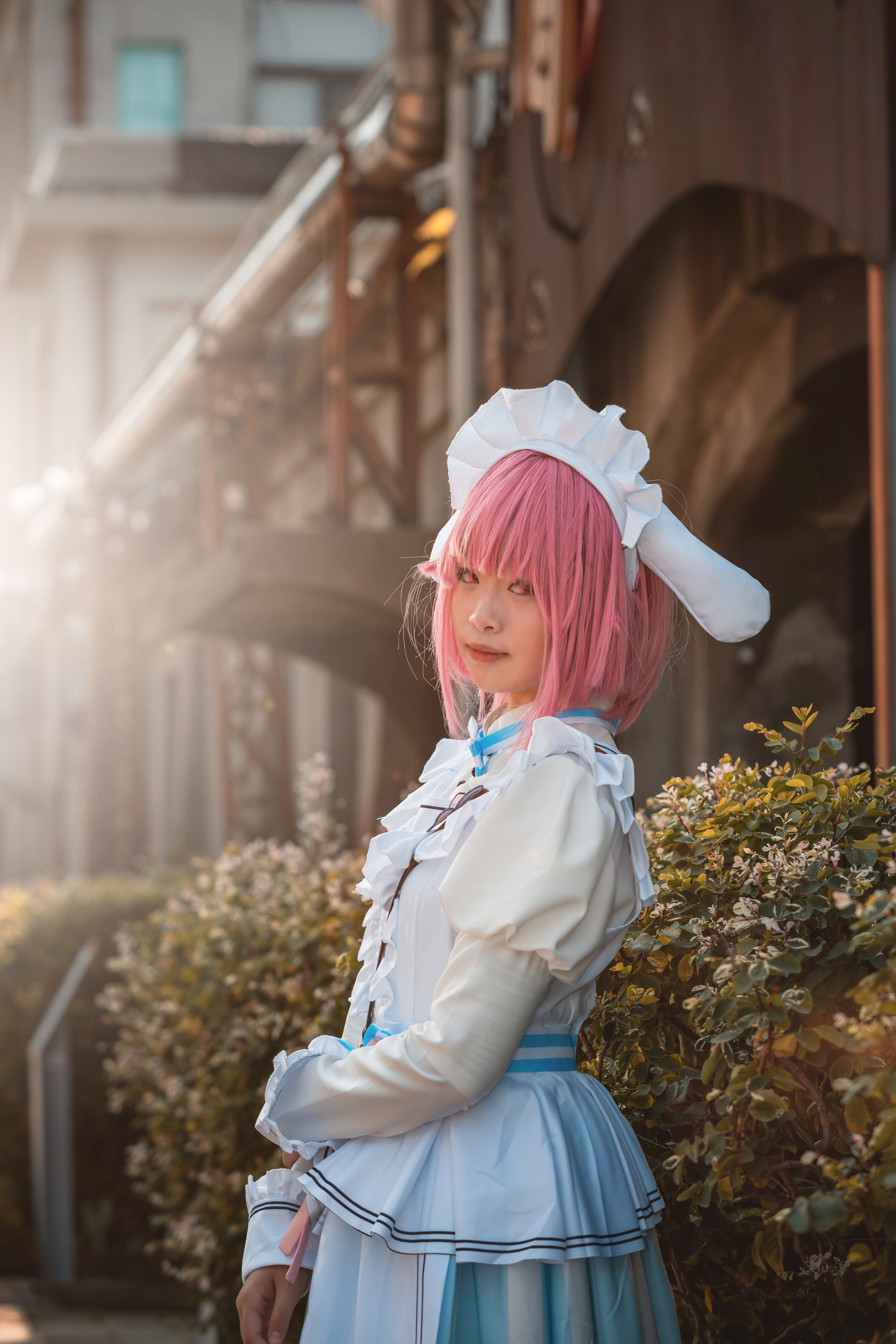 Cosplayer in detailed pink wig and costume posing outdoors during the day.