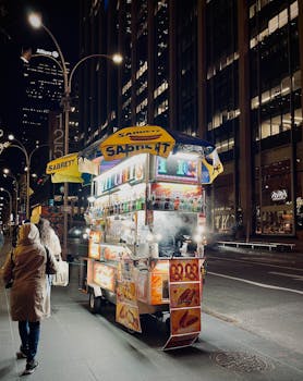 Brightly lit hot dog cart on New York City street at night with skyscrapers.