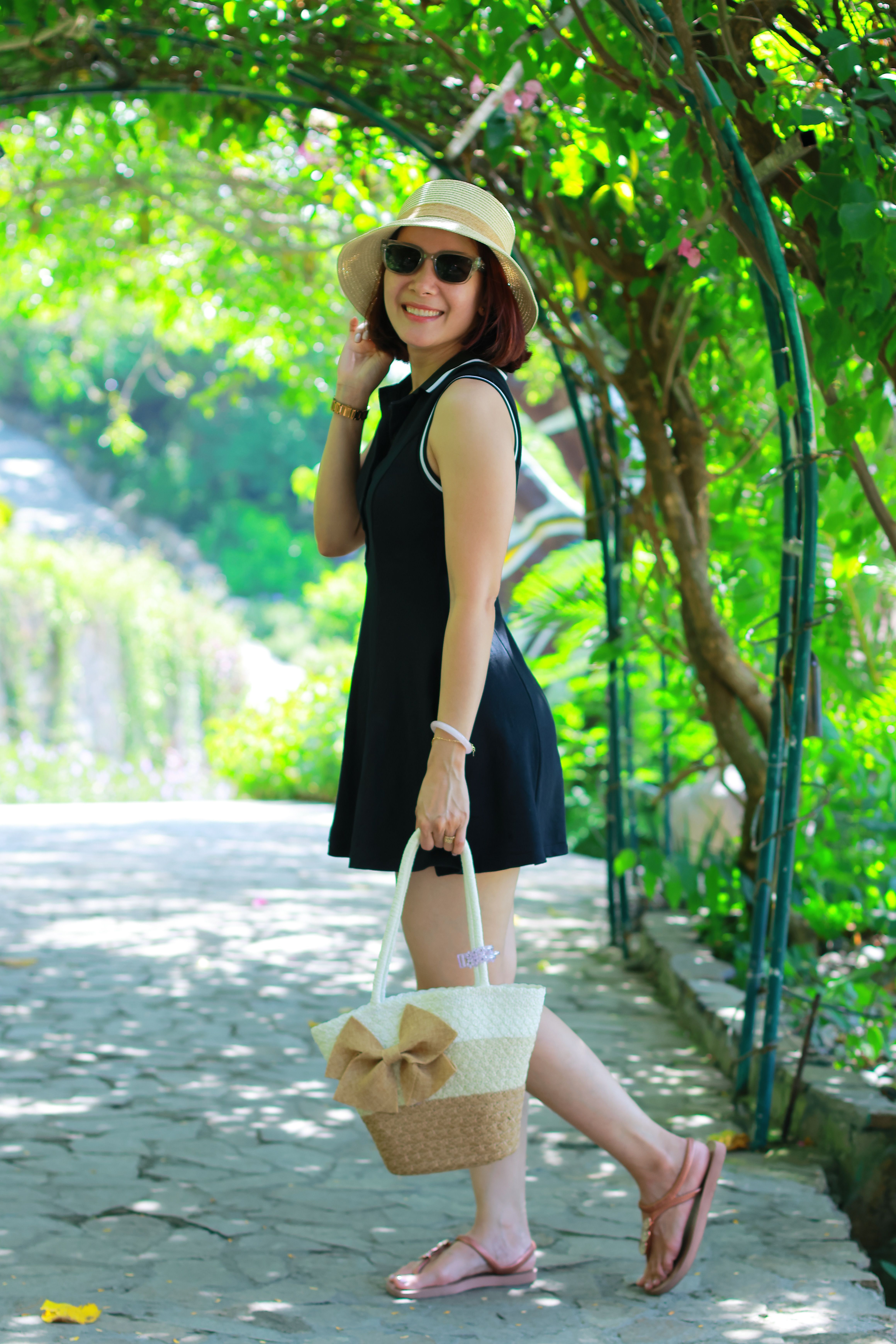 Smiling woman in summer dress and hat strolling through lush garden archway.