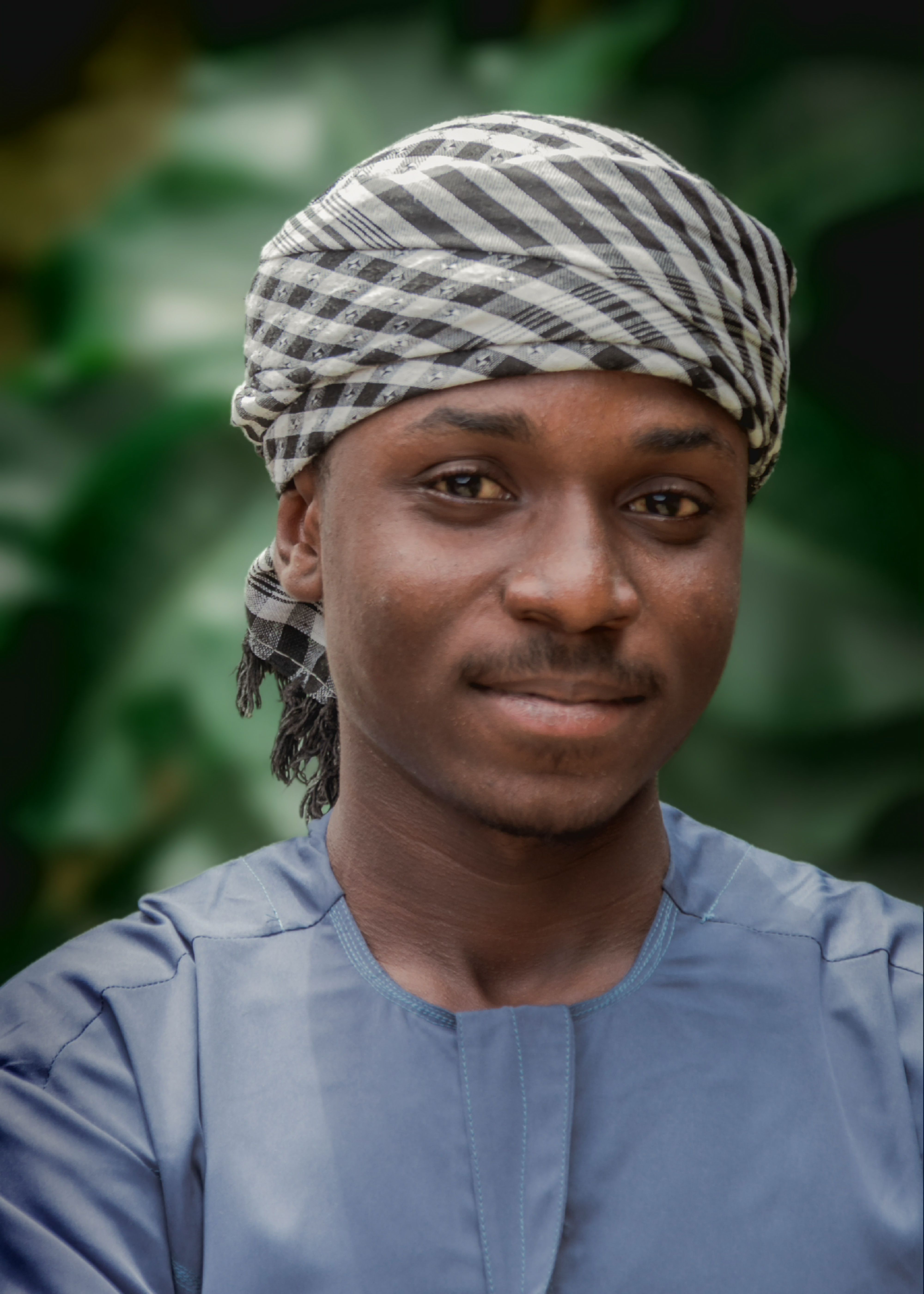 A close-up portrait of a man wearing a striped headwrap and blue traditional attire, smiling outdoors.