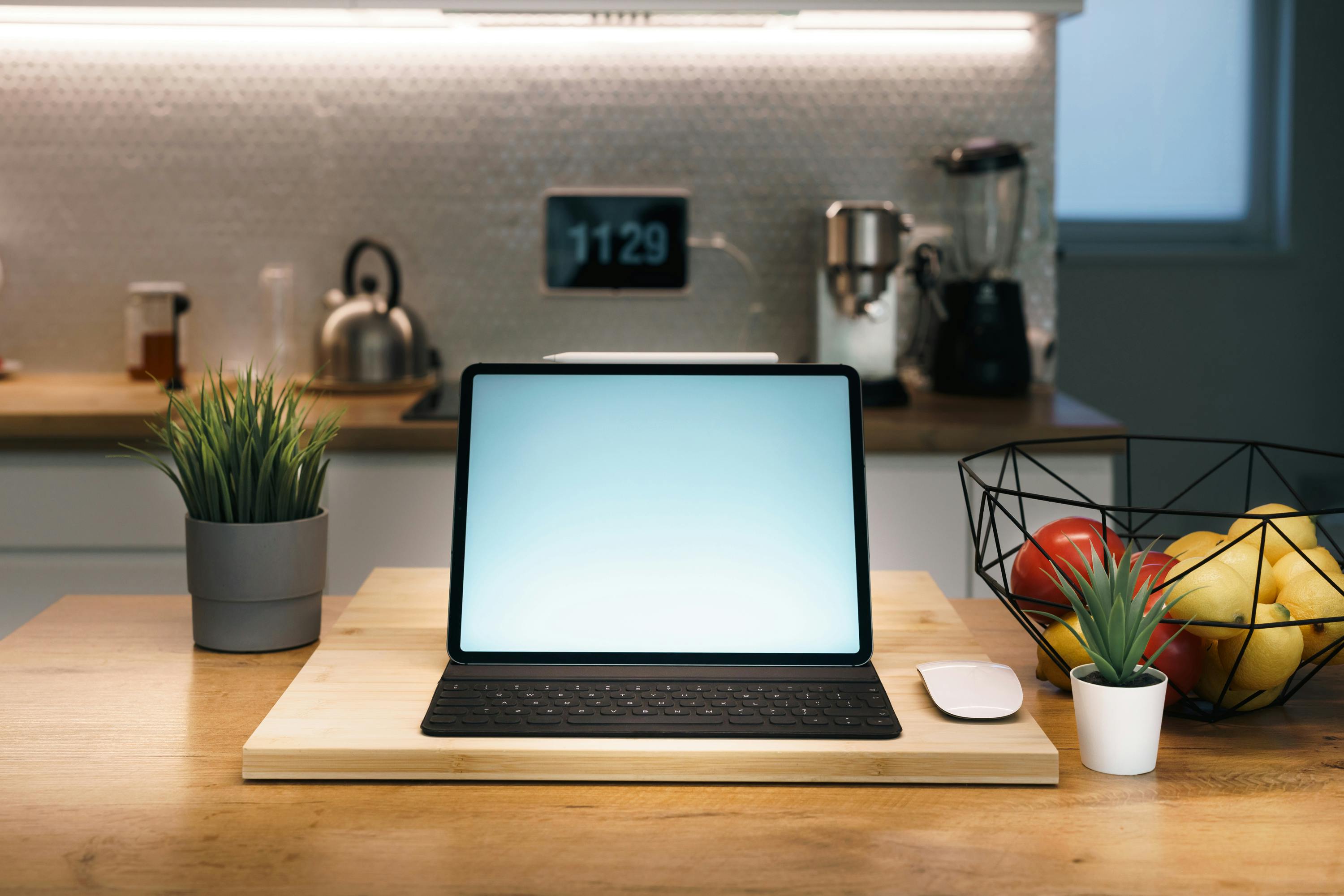 Cozy home workspace with digital tablet on a kitchen counter, surrounded by plants and fruit bowl, perfect for remote work.