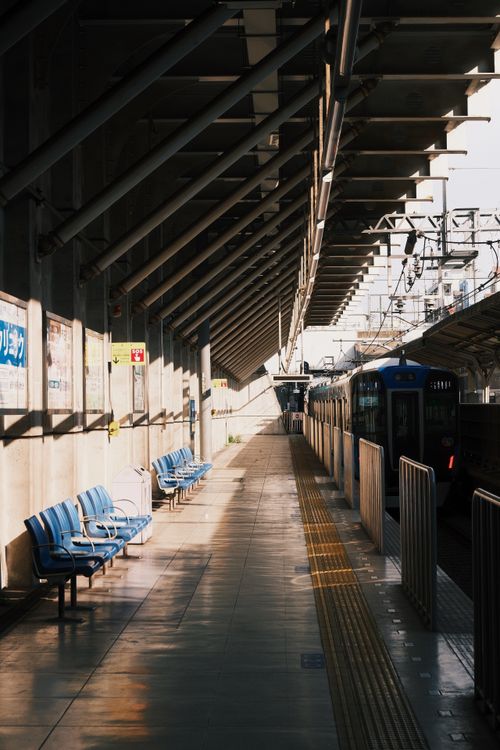 Free Sunlit train station platform with empty blue seats and arriving train, urban setting. Stock Photo
