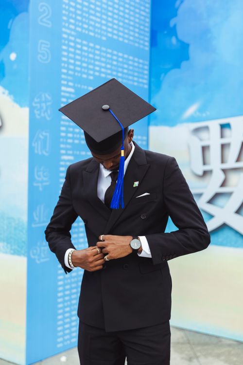 Free A young graduate, dressed in a suit and graduation cap, adjusting his attire outdoors. Stock Photo