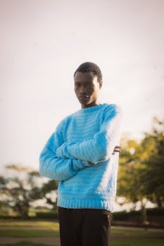 Young man with blue sweater poses confidently outdoors in natural light.