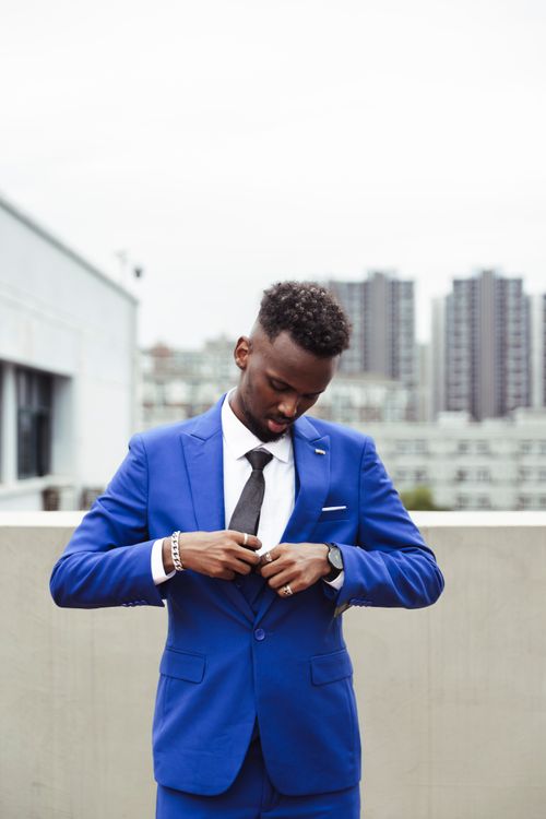 Free Man in bright blue suit adjusting his tie on an urban rooftop, showcasing modern fashion. Stock Photo