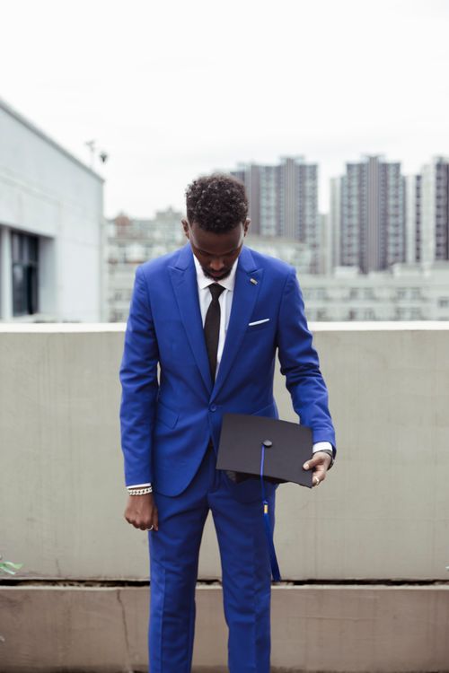 Free Adult male graduate in blue suit holding cap on a rooftop with city skyline backdrop. Stock Photo