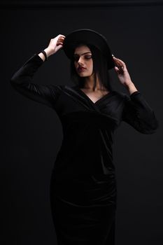 Fashionable woman posing elegantly in dark studio, wearing a black dress and stylish hat.