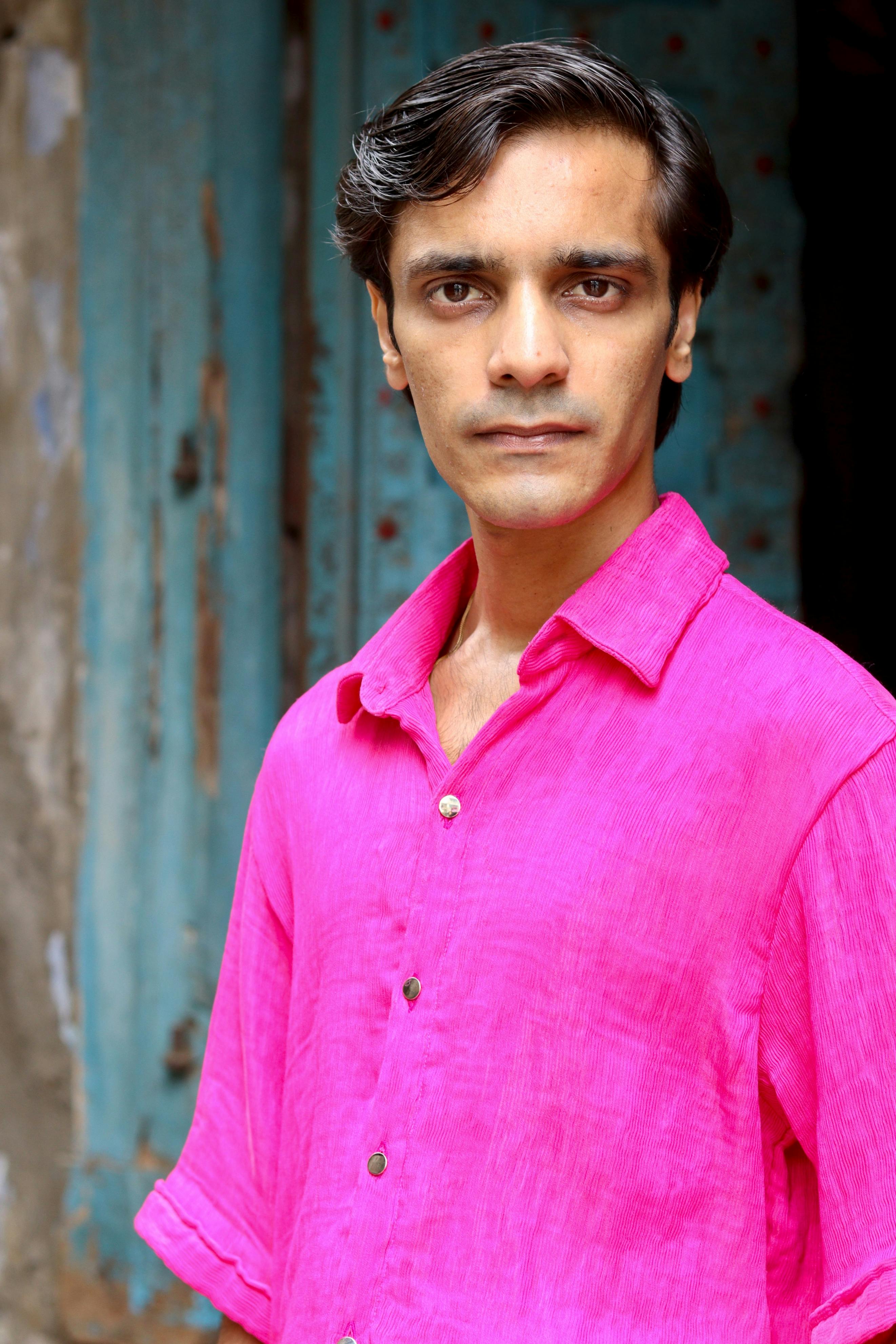 Confident man in pink shirt standing against a rustic blue door, showcasing bold urban style.