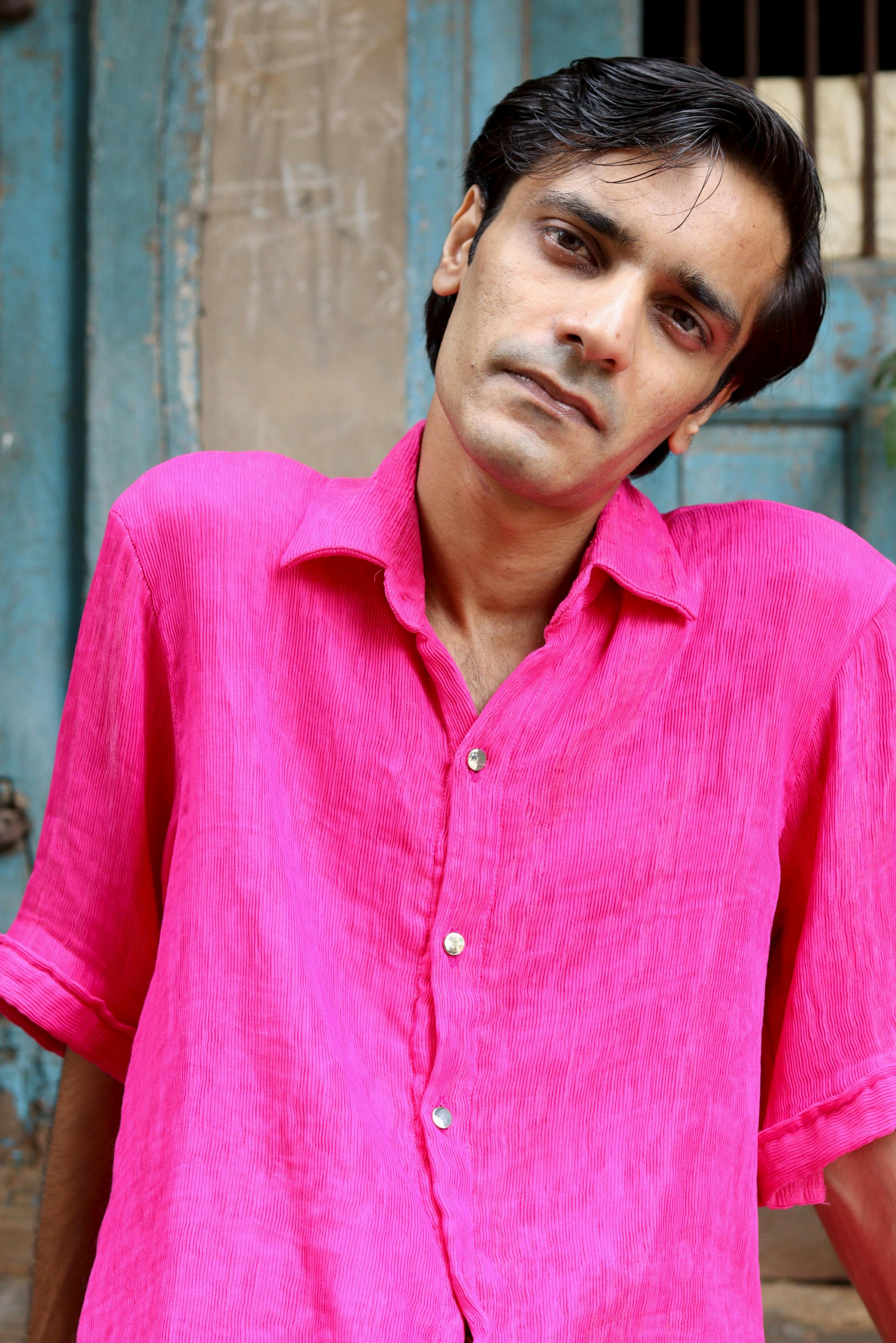 Portrait of a man in a bright pink shirt leaning against a rustic wall.