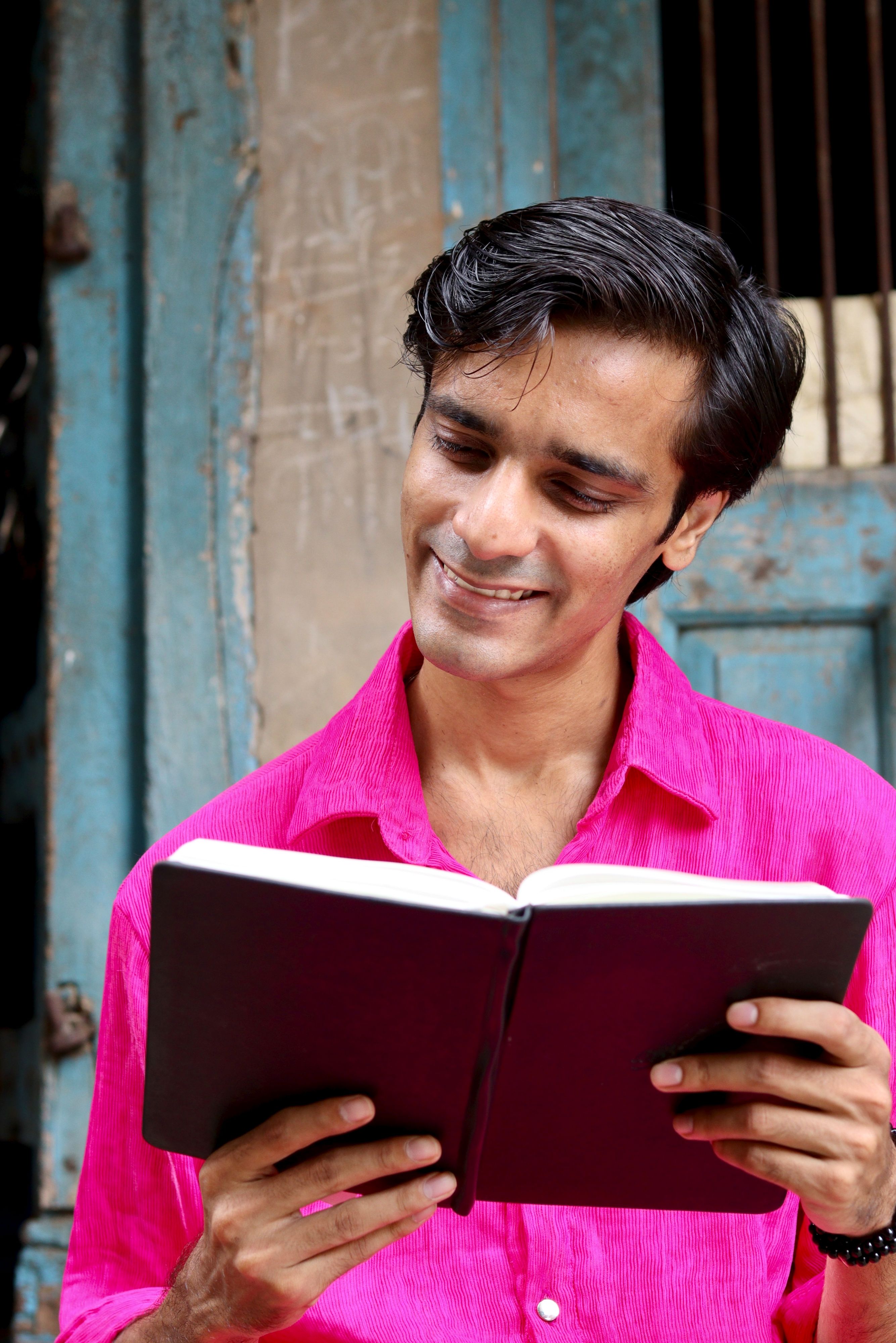 Man smiling while reading a book outdoors against a rustic background.