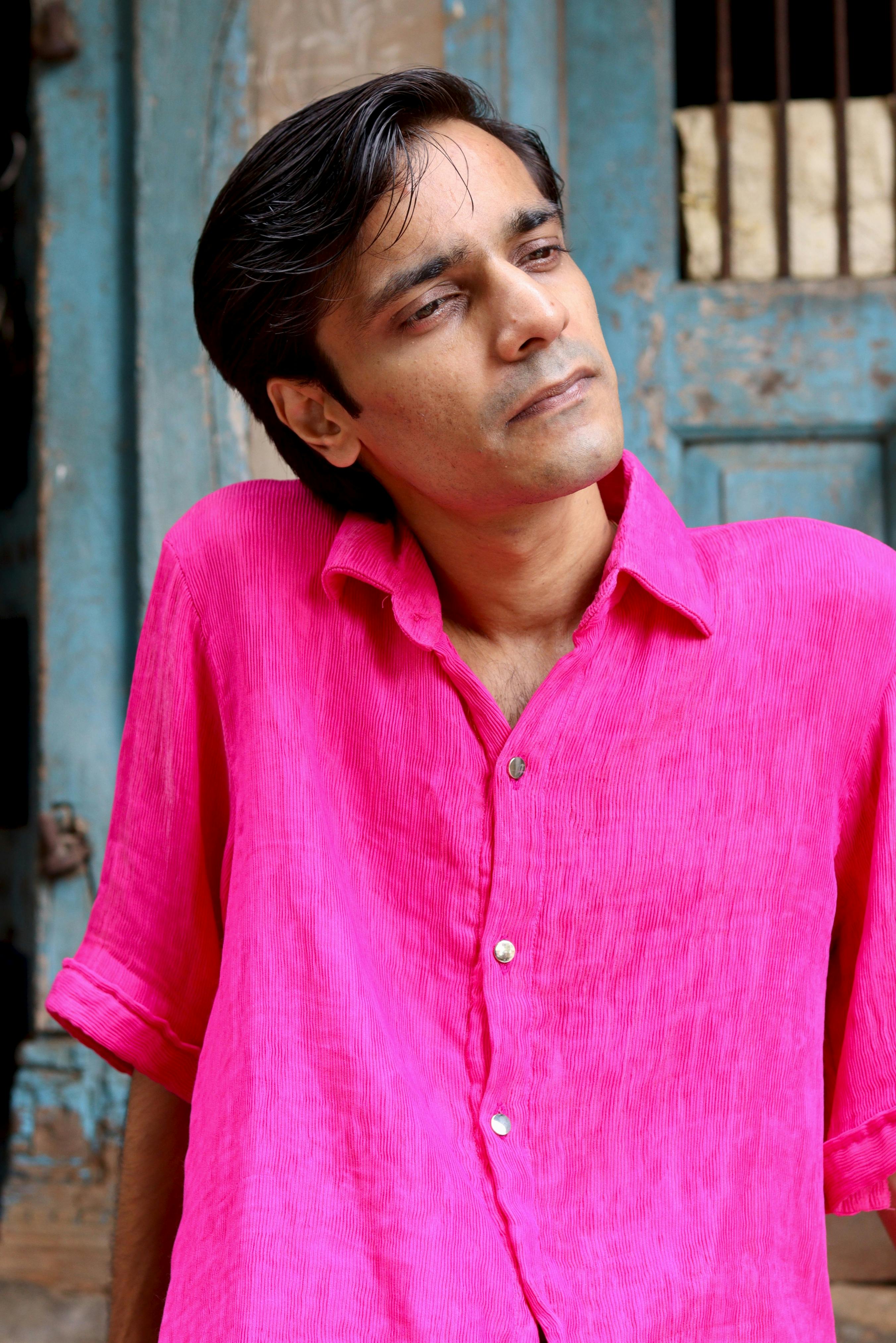 A young man poses thoughtfully in a bright pink shirt in an outdoor setting.