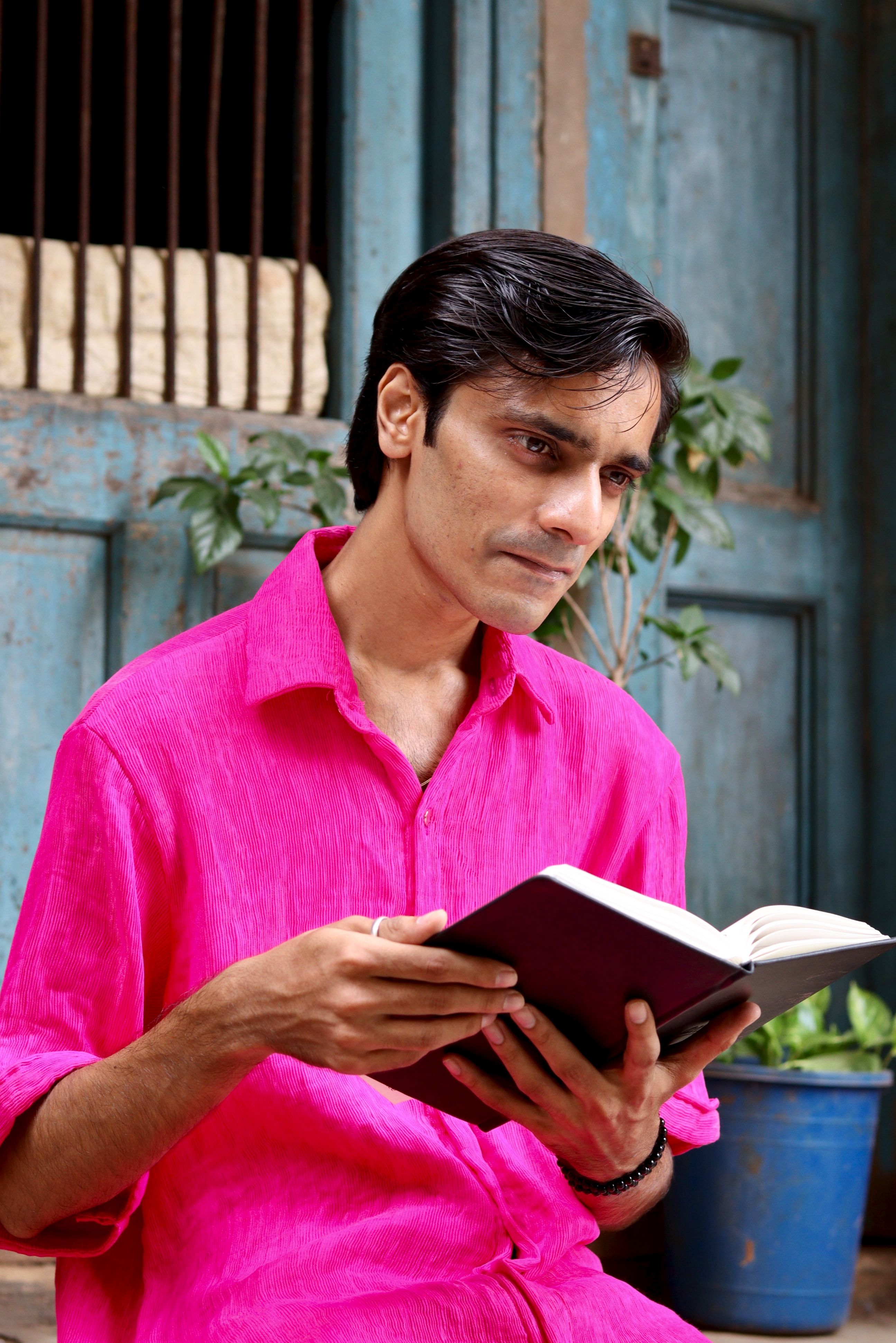 Portrait of a man in a bright pink shirt reading a book outdoors by a rustic setting.