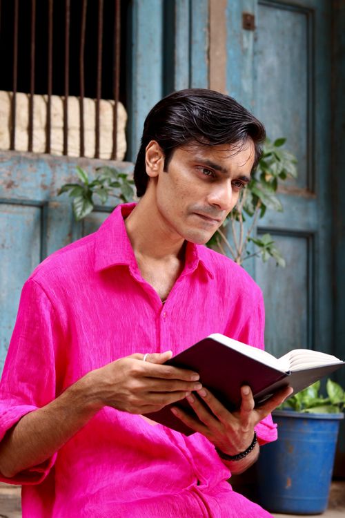 Free Portrait of a man in a bright pink shirt reading a book outdoors by a rustic setting. Stock Photo