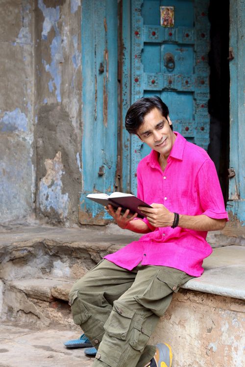 Free Smiling man in pink shirt reads book outdoors near rustic blue door. Stock Photo