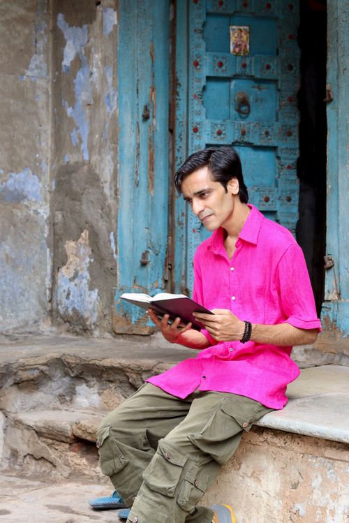 Free A young man in a vibrant pink shirt reads a book outdoors against a rustic blue door. Stock Photo