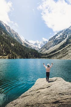 A young child stands on a rock, arms raised, overlooking a tranquil mountain lake and snow-capped peaks.