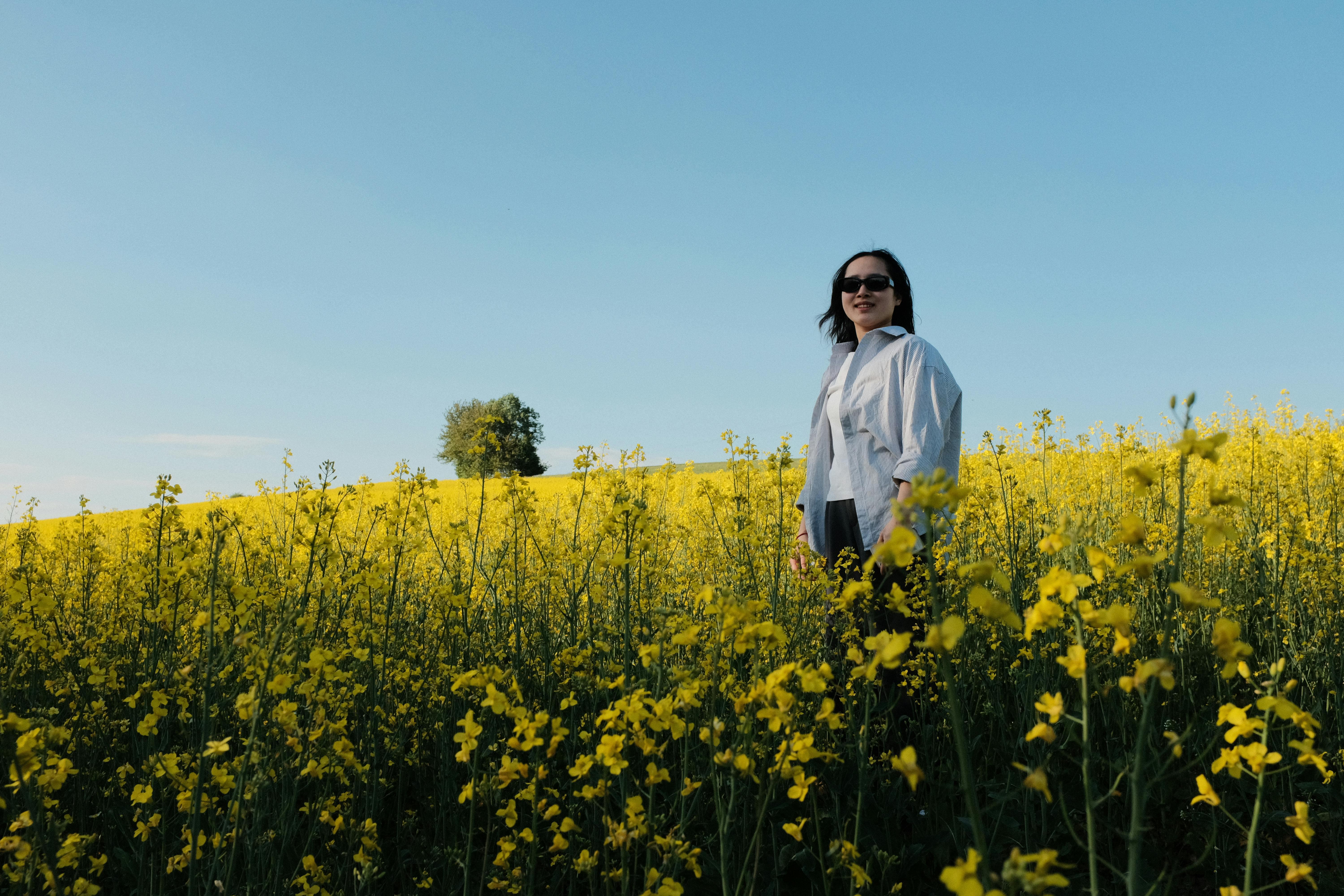 A woman stands in a vibrant yellow field under a clear blue sky, embracing nature's beauty.
