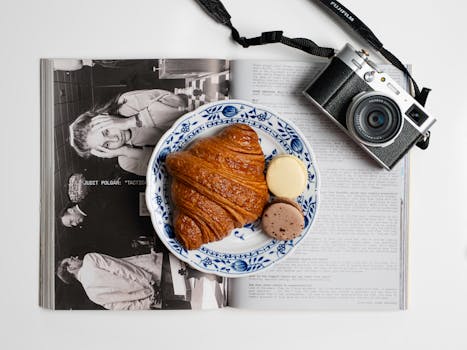 Artistic flat lay of a croissant and macarons on a fashion magazine with a camera, perfect for food photography.