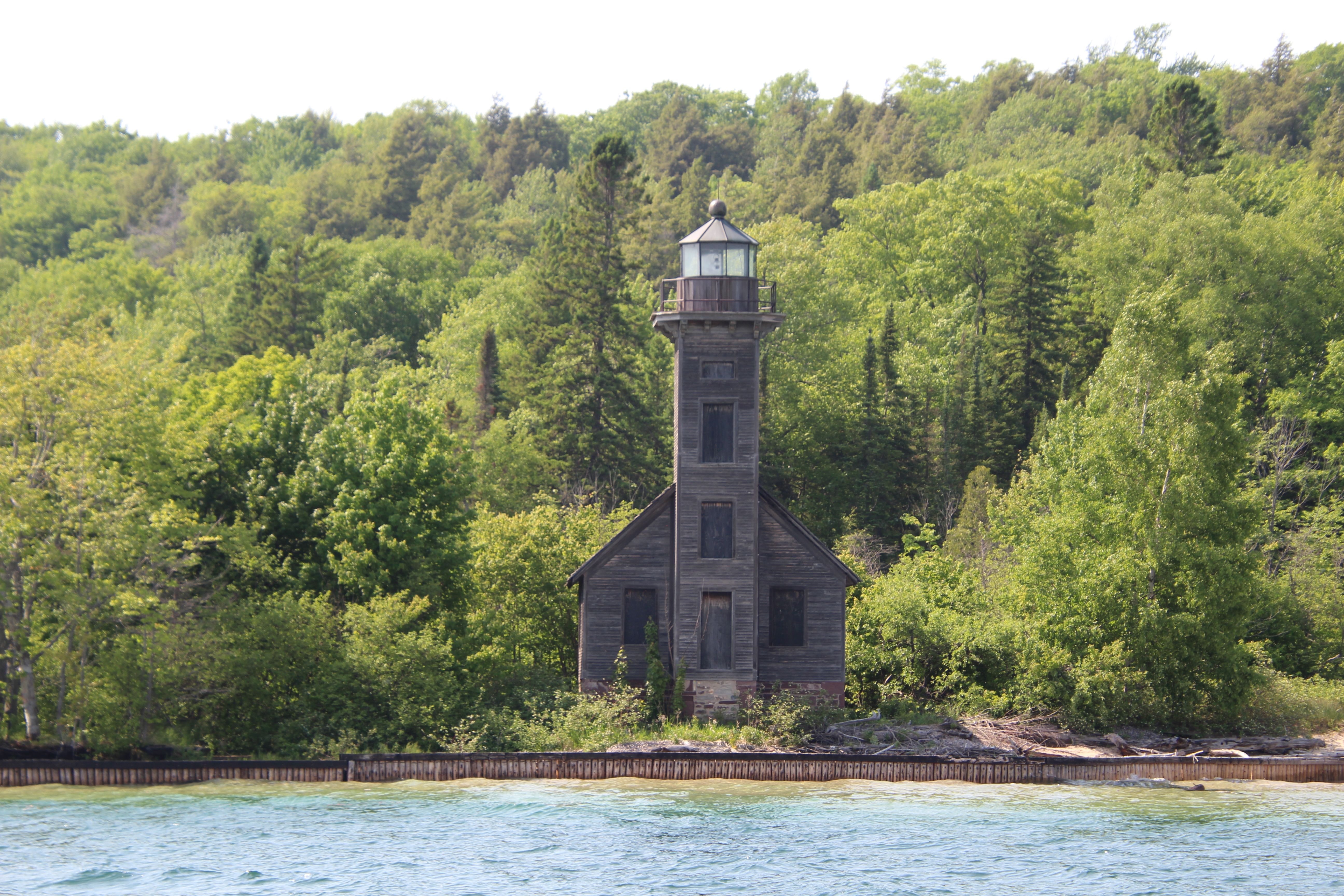Old wooden lighthouse surrounded by dense forest and calm waters in a tranquil natural setting.