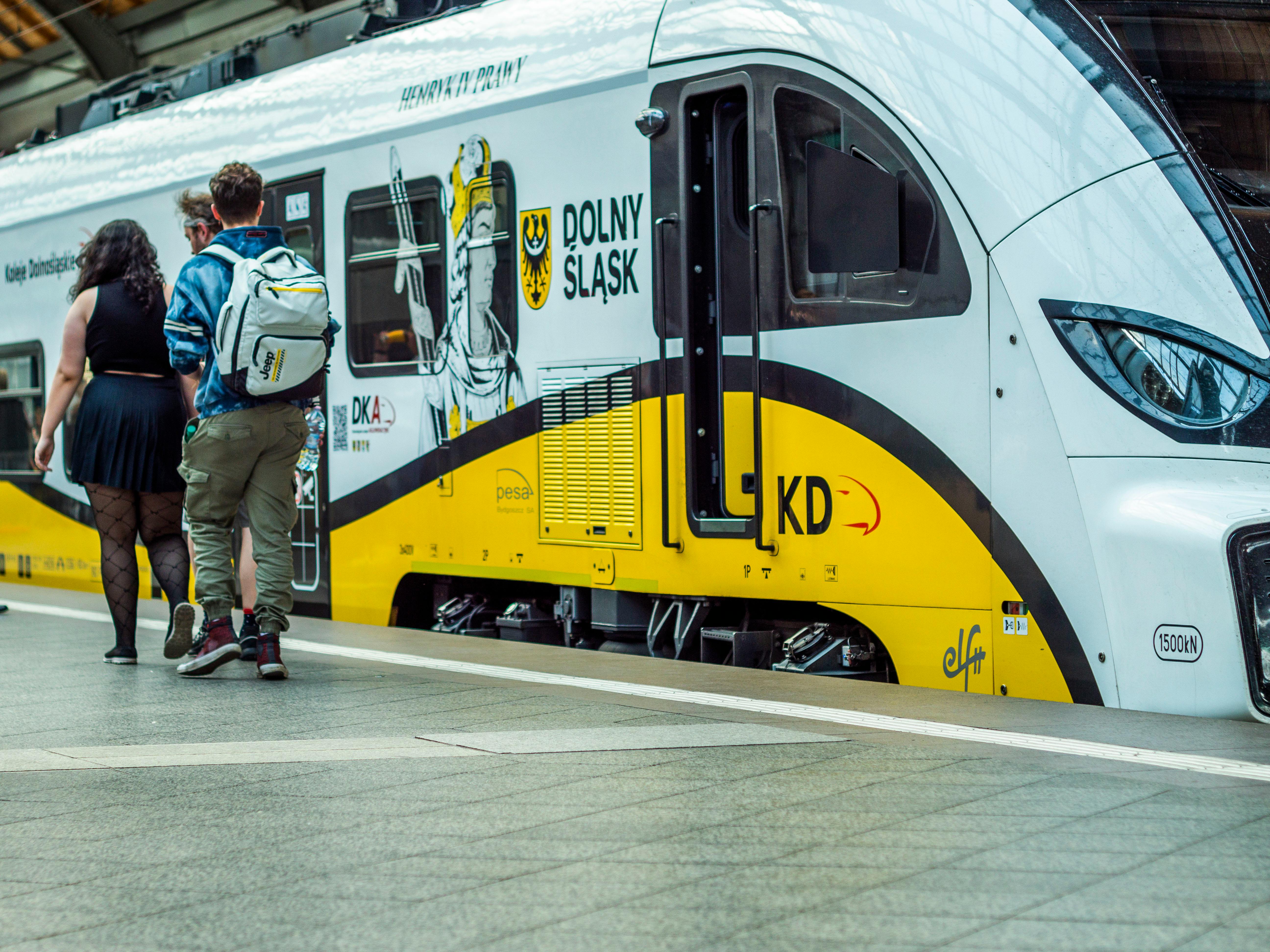 Passengers with backpacks walk along a platform beside a yellow and white train at a station