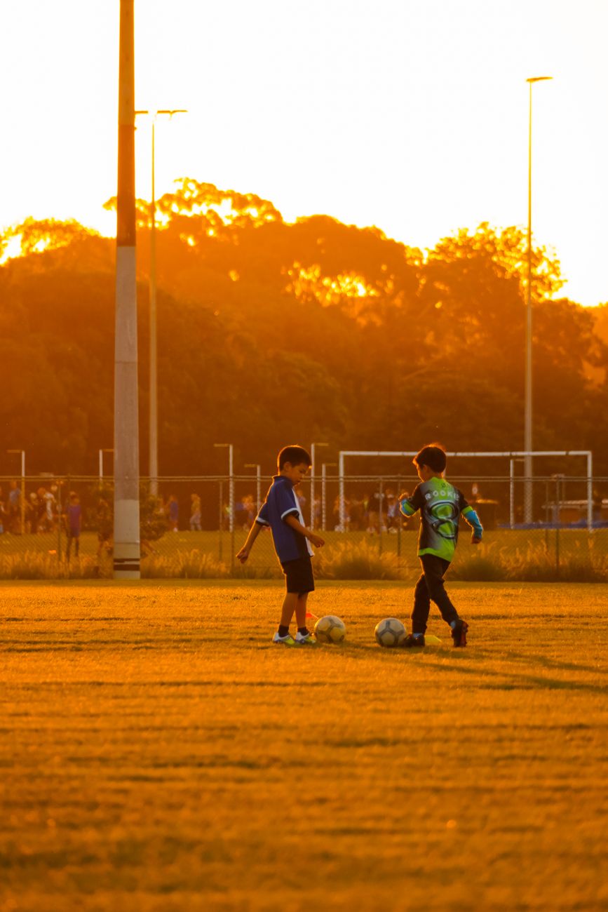 Two kids playing soccer on a field during sunset, vibrant golden hues.