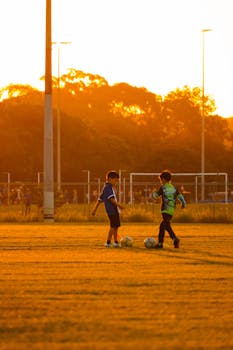 Two kids playing soccer on a field during sunset, vibrant golden hues.
