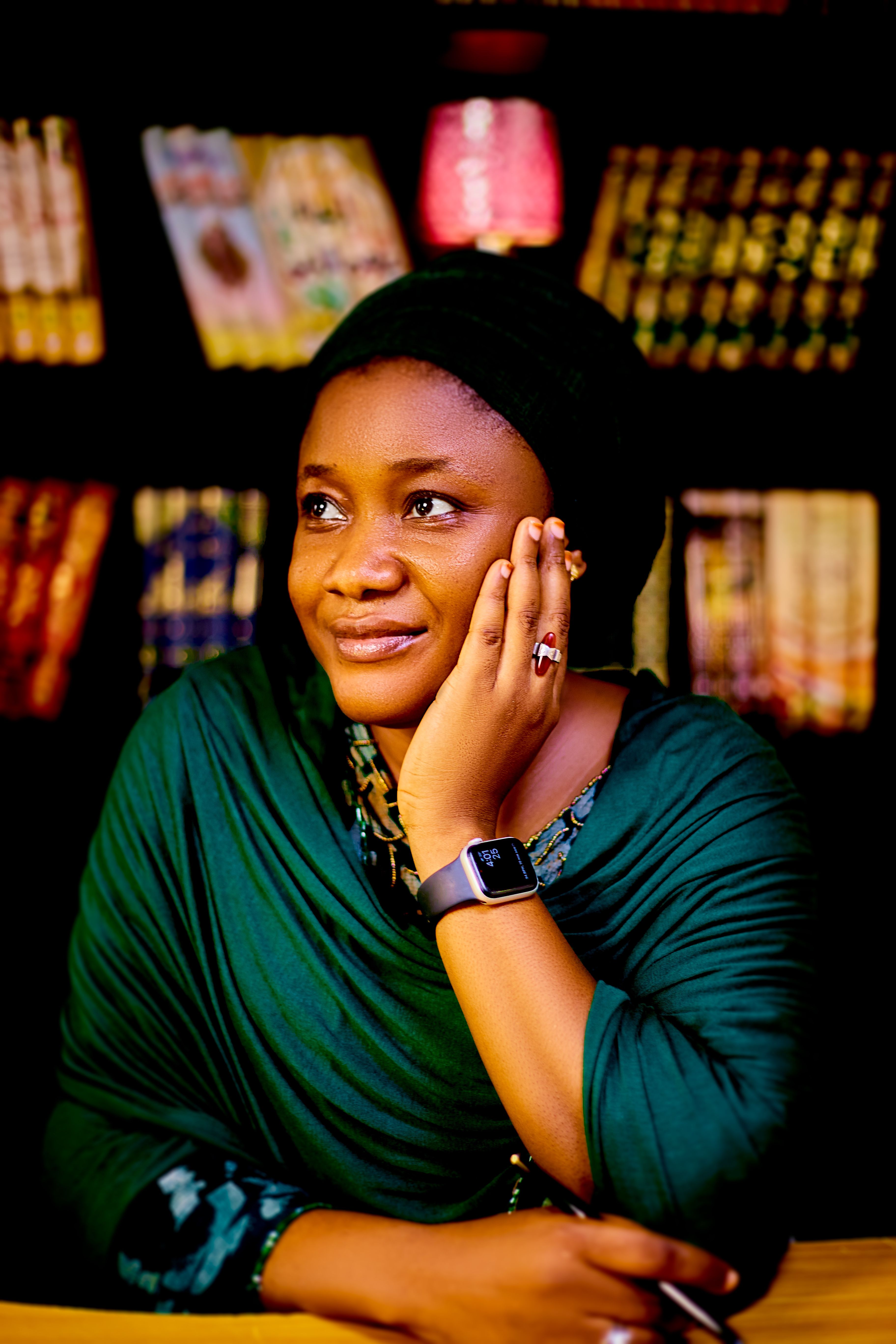 A thoughtful woman in a green shawl sits in a warmly lit library, smiling gently.