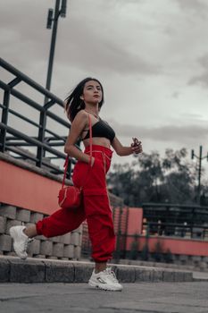 Fashionable young woman wearing a trendy red outfit walking confidently outdoors on a cloudy day.