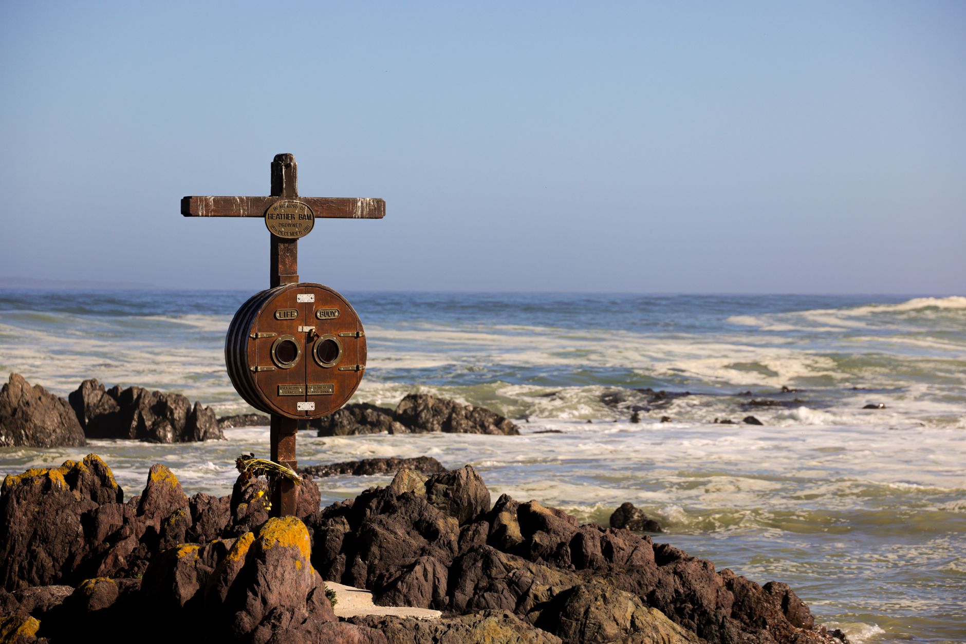 Wooden coastal warning sign on rocky beach with ocean waves in background.