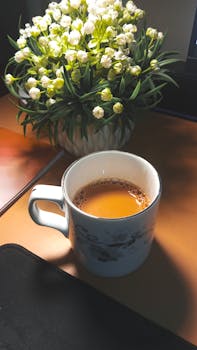 A warm cup of tea sits peacefully on a desk beside a white floral arrangement, capturing a calm moment.