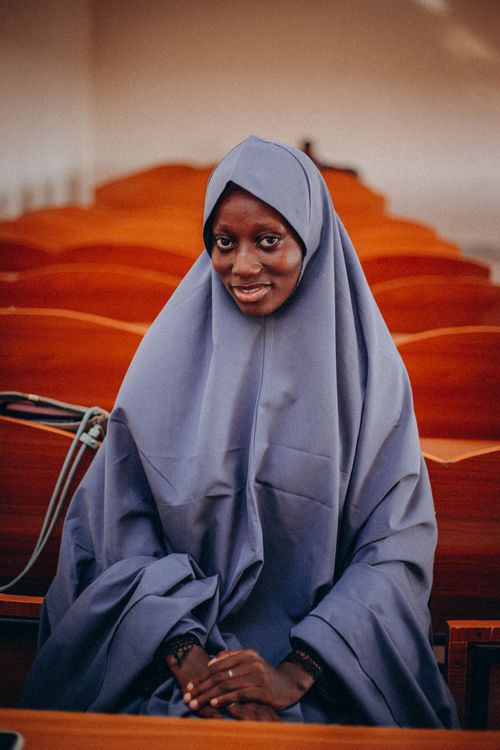 Free Smiling Muslim woman wearing hijab in an indoor prayer hall with wooden seats. Stock Photo