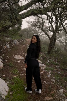 A woman in black clothing walks on a misty forest trail surrounded by rocky terrain and moss-covered trees.
