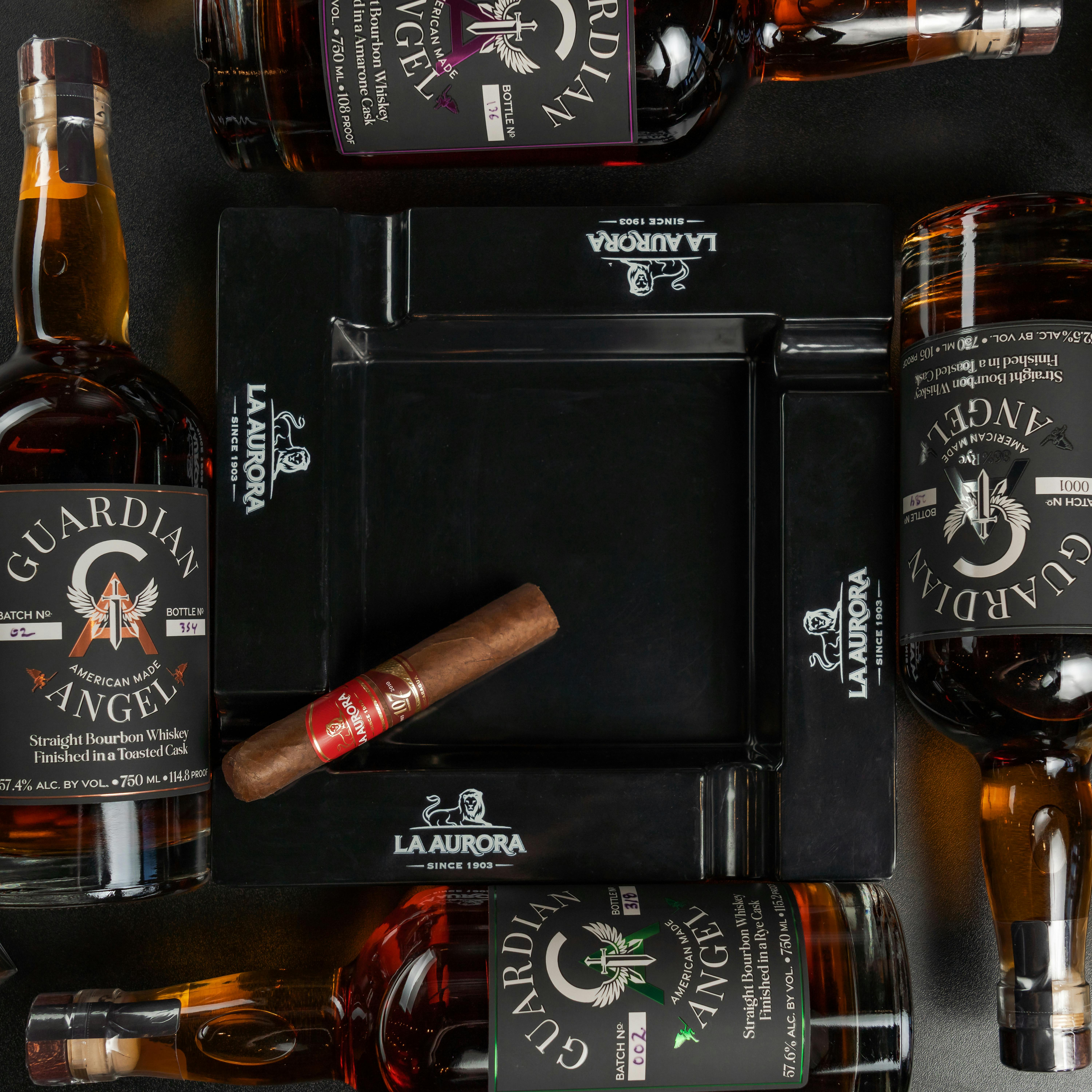 Flat lay of Guardian Angel whiskey bottles, a cigar, and La Aurora ashtray.