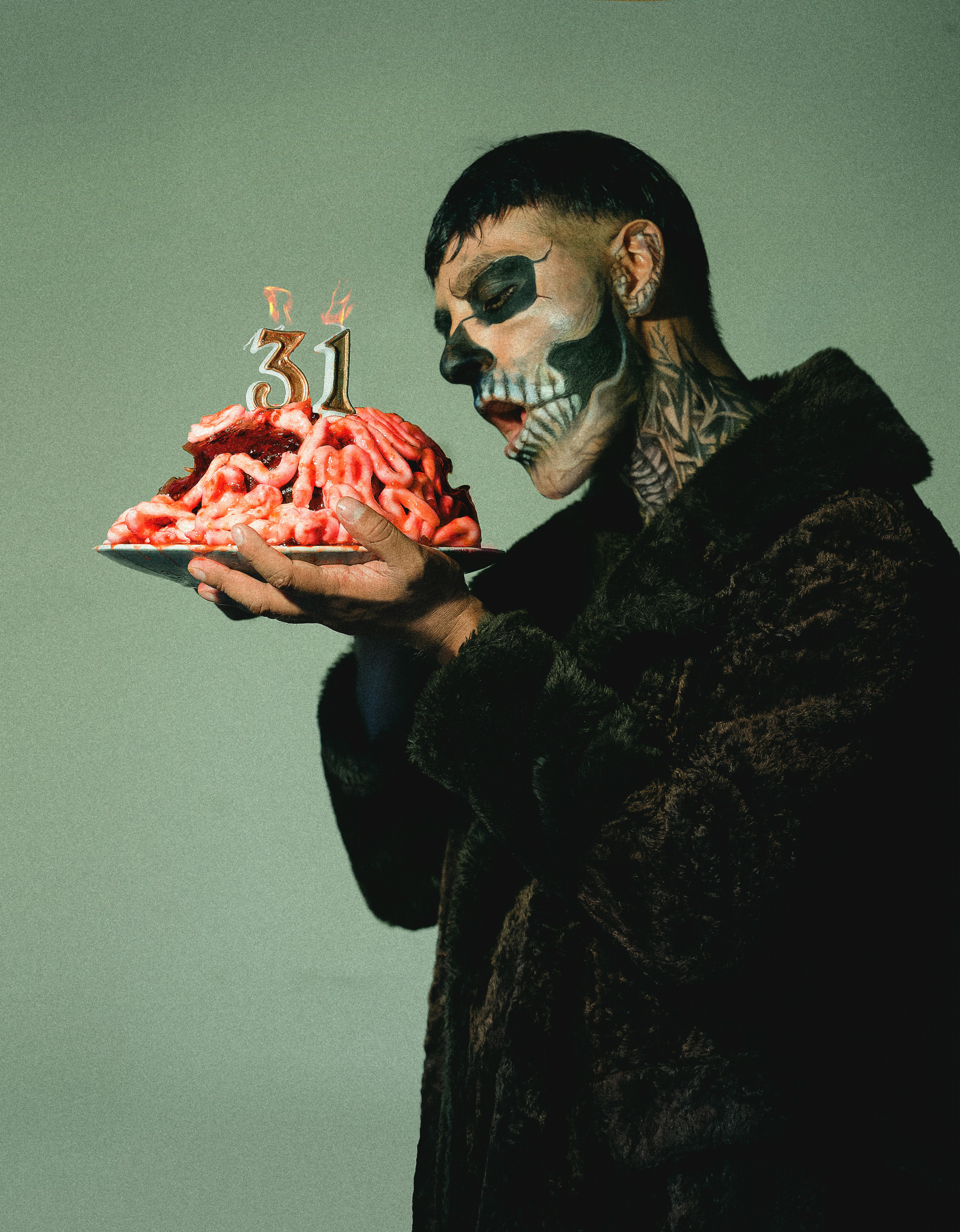 A person with intricate skull makeup holds a cake resembling a brain with '31' candles, demonstrating artistic creativity.