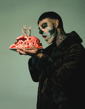 A person with intricate skull makeup holds a cake resembling a brain with '31' candles, demonstrating artistic creativity.