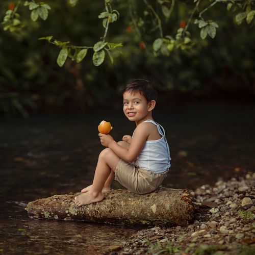 Free A happy child sitting on a log by a river, enjoying a mango in Tingo María, Peru. Stock Photo