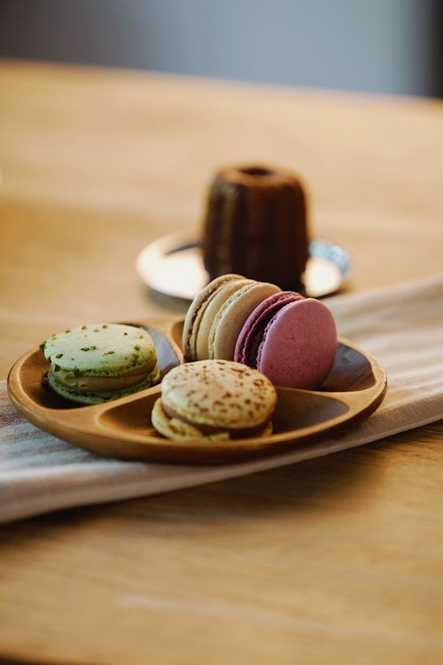 Free A selection of colorful macarons on a wooden plate with a pastry in the background. Stock Photo
