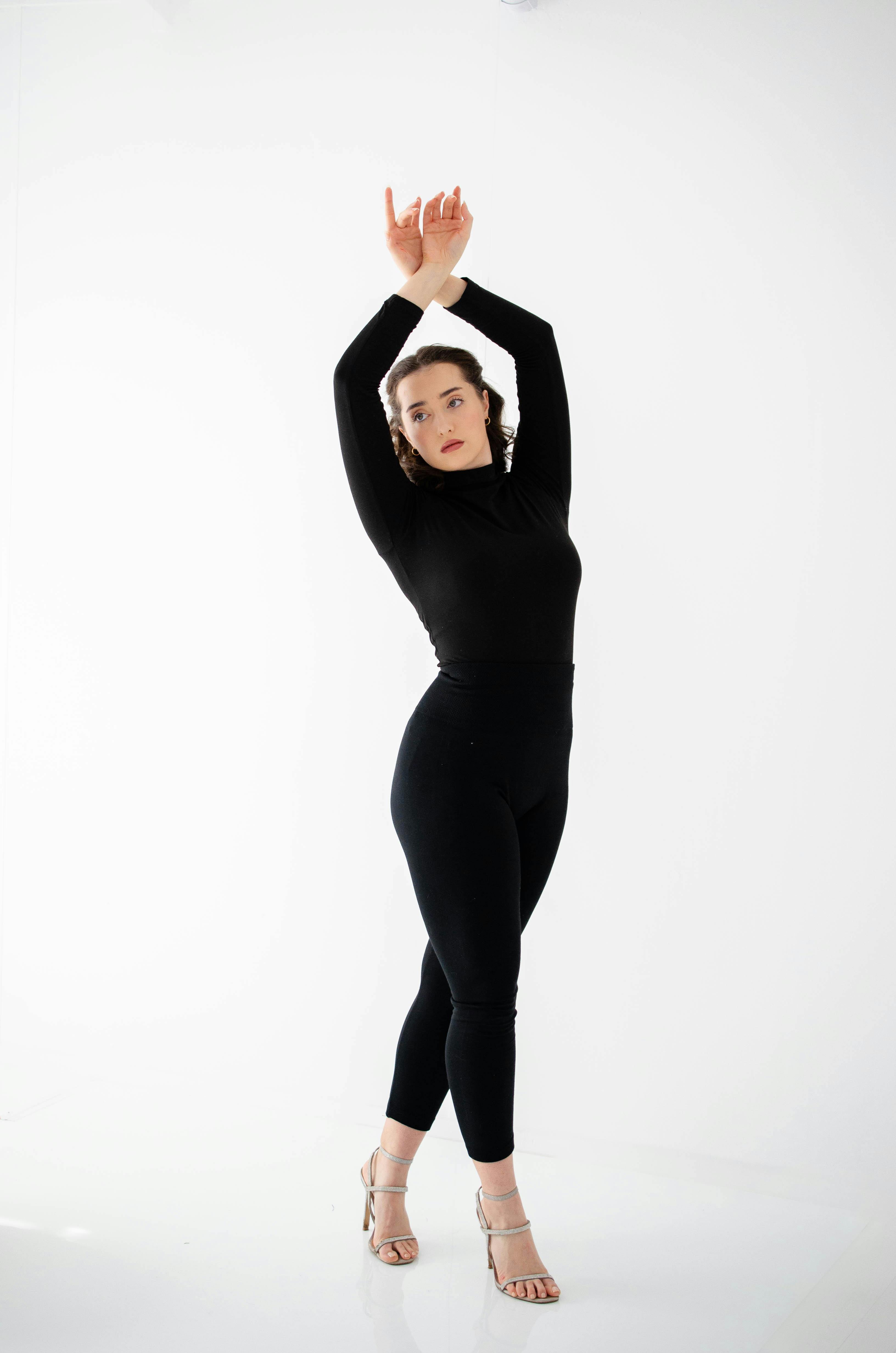 Woman in elegant black outfit striking a pose in a minimalist white space.