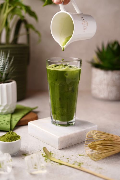 De franc Un got alt de cafè amb llet matcha gelat amb nata, envoltat de matcha i plantes. Foto d'estoc
