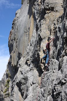 Man climbing steep rock wall with ropes on a sunny day, showcasing outdoor adventure and skill.