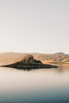 A serene island surrounded by water, set against a picturesque mountain backdrop during sunset.